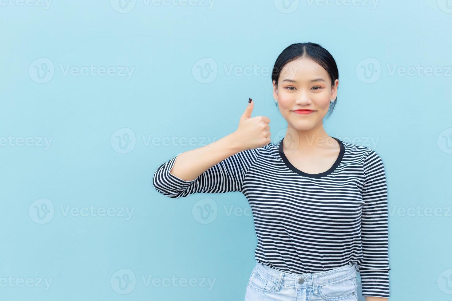 Portrait of cute Asian Thai girl student looking friendly is standing smiling happily and confidently successful showing thump up for communication as good to present something on a blue background. photo