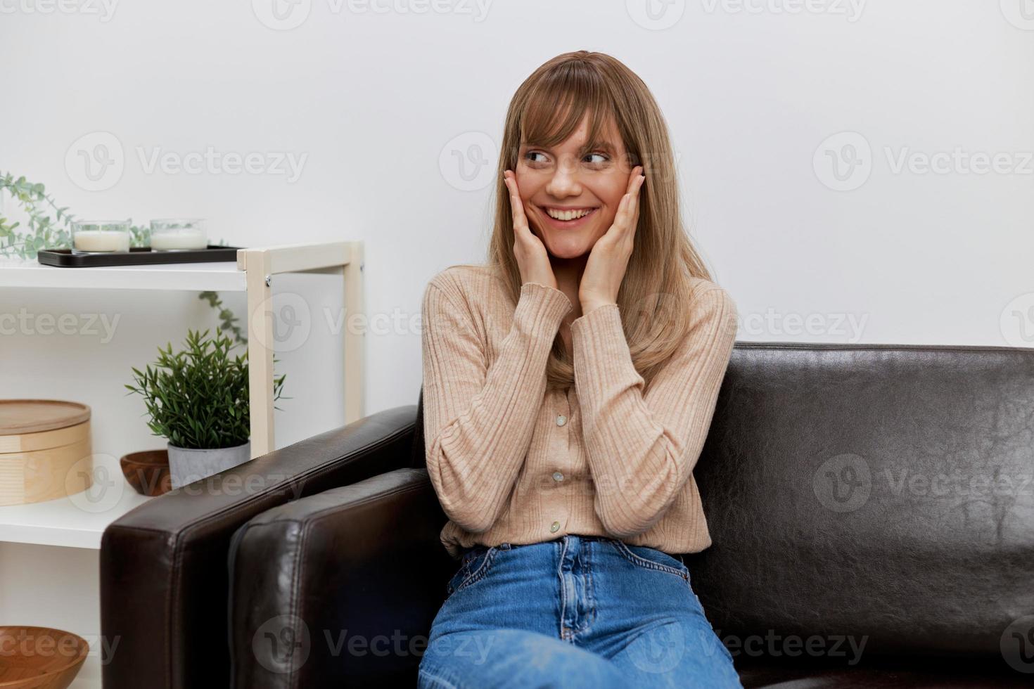 alegre gracioso rubia dama en casual suéter toques las mejillas mira aparte sentado en sofá a moderno vivo habitación interior. hembra cliente tener encogimiento cita en psicológico ayuda oficina. Copiar espacio foto