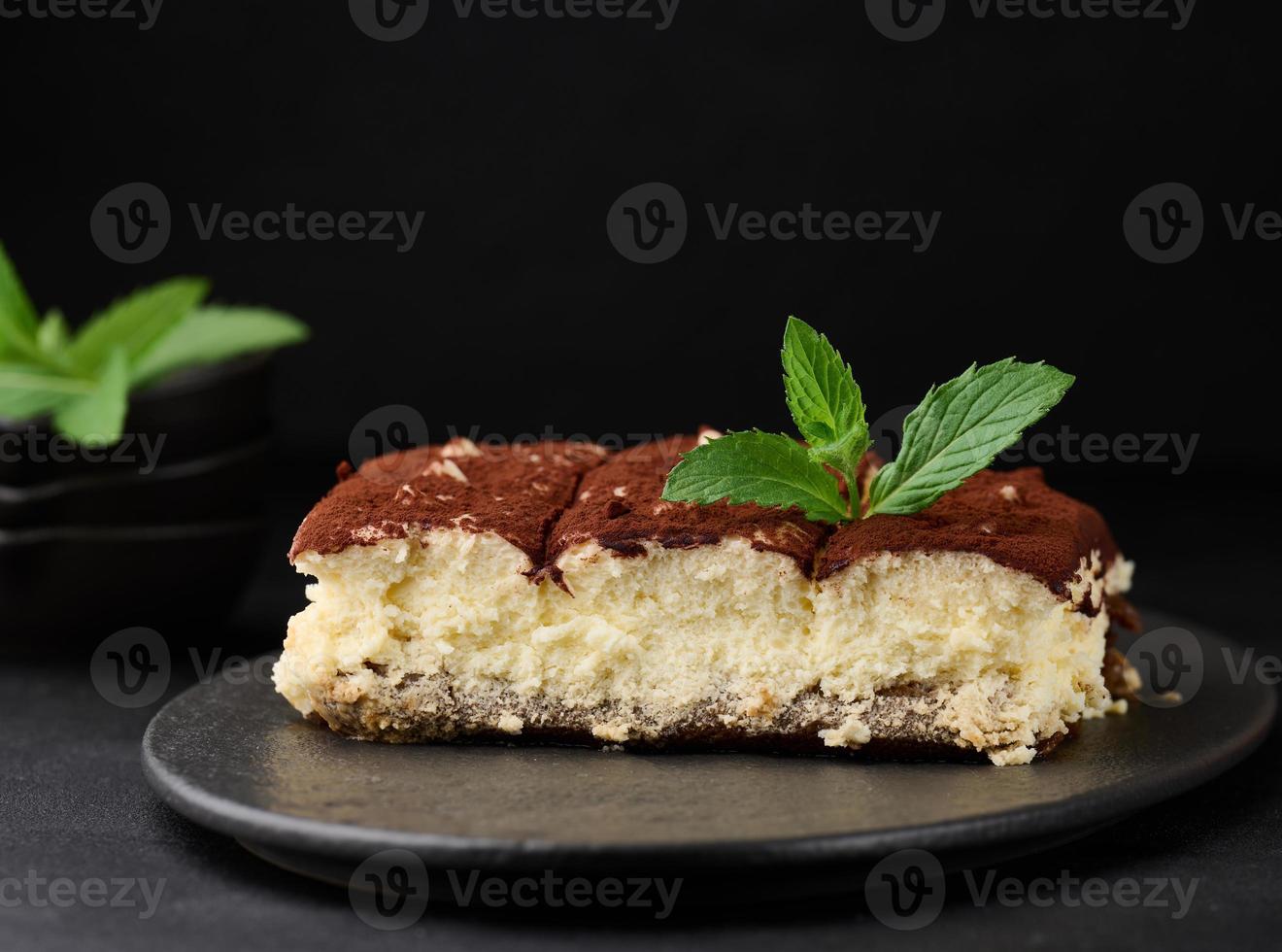 A piece of tiramisu sprinkled with cocoa, on top of a sprig of fresh mint on a black background photo