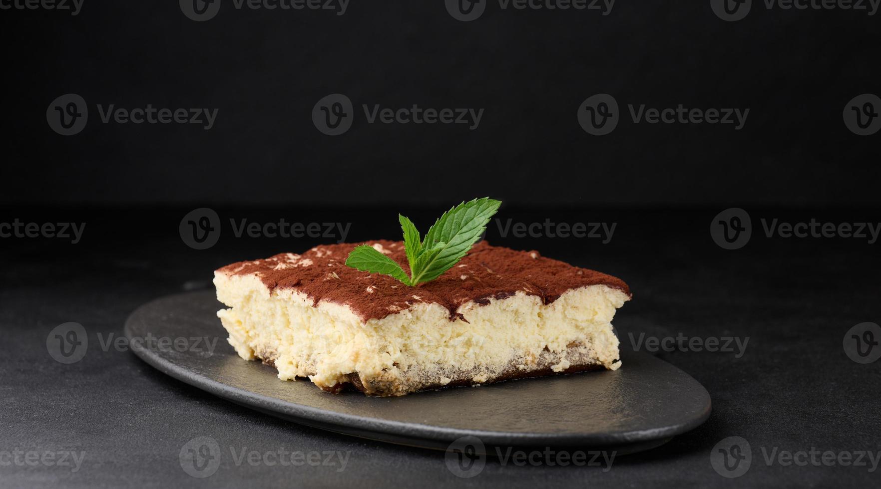 A piece of tiramisu sprinkled with cocoa, on top of a sprig of fresh mint on a black background photo