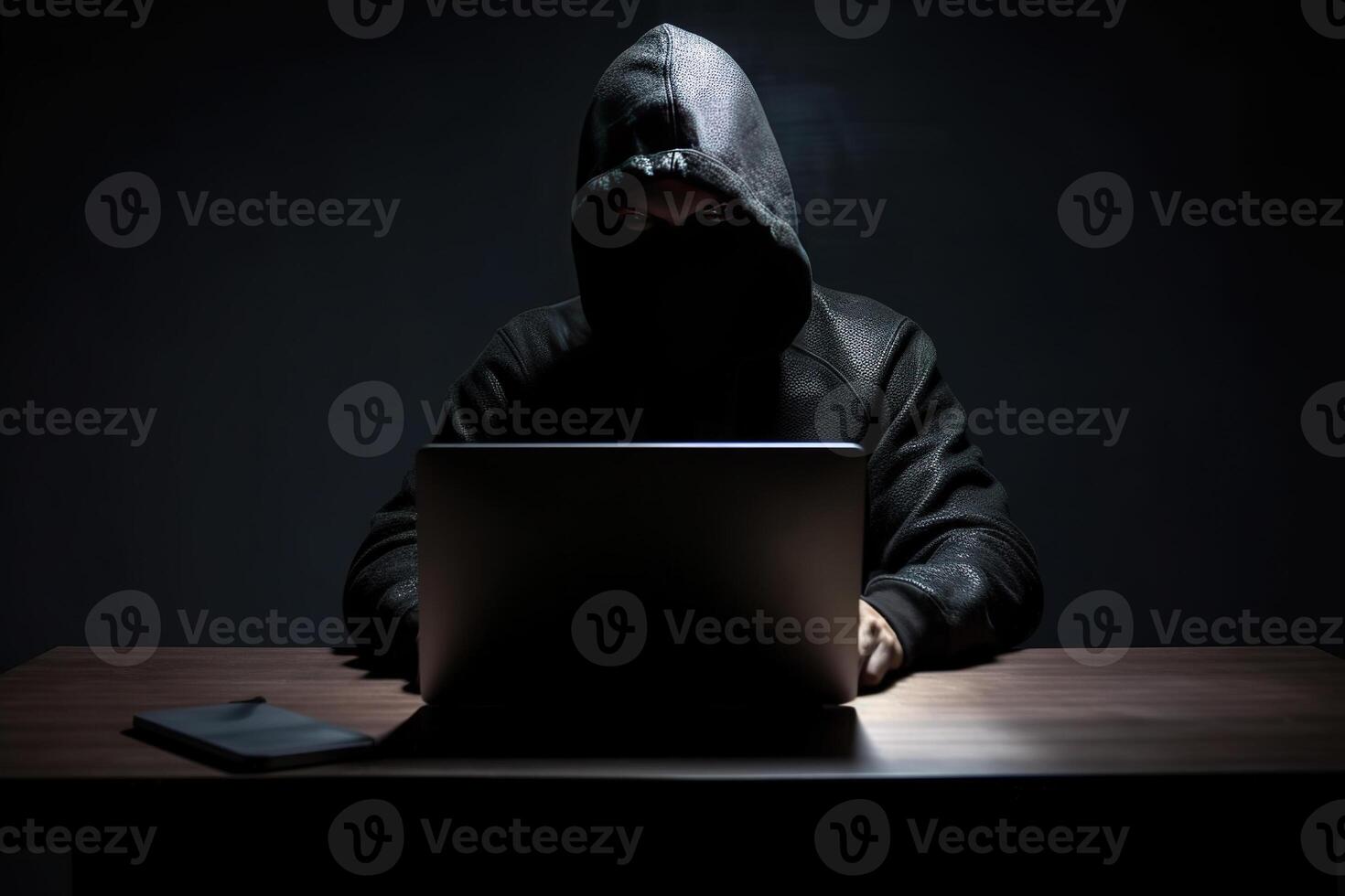 Hacker wearing black hoodie using laptop computer while working on dark background. photo