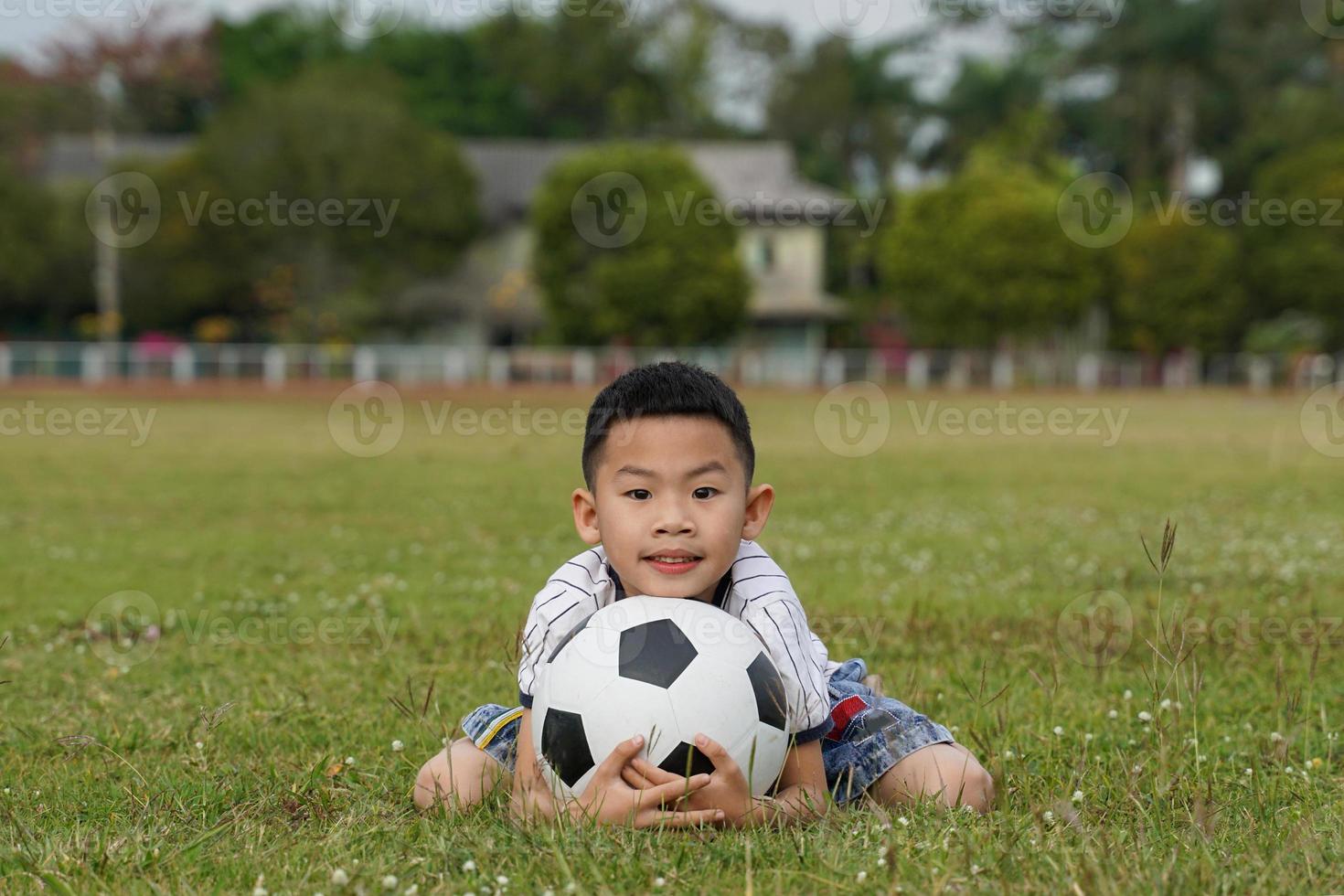 Asian boy sits with his chin on a soccer ball in the grass, in the concept of outdoor activities, sports, playgrounds, leisure activities. Soft and selective focus. photo