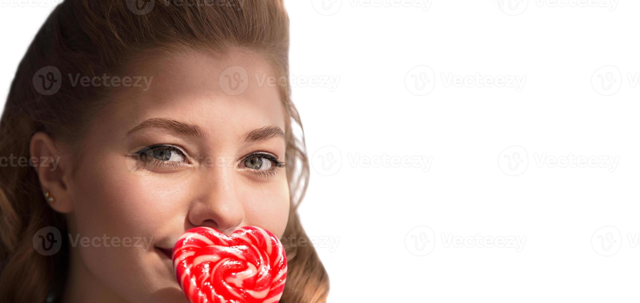 The face of a girl with candy on a stick in the form of a heart on a white background. Sweet woman. photo