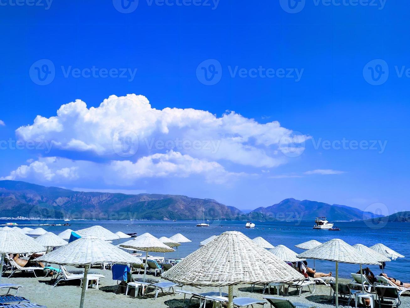 a scene with the sea, mountains, blue sky, and straw umbrellas providing shade from the sun photo