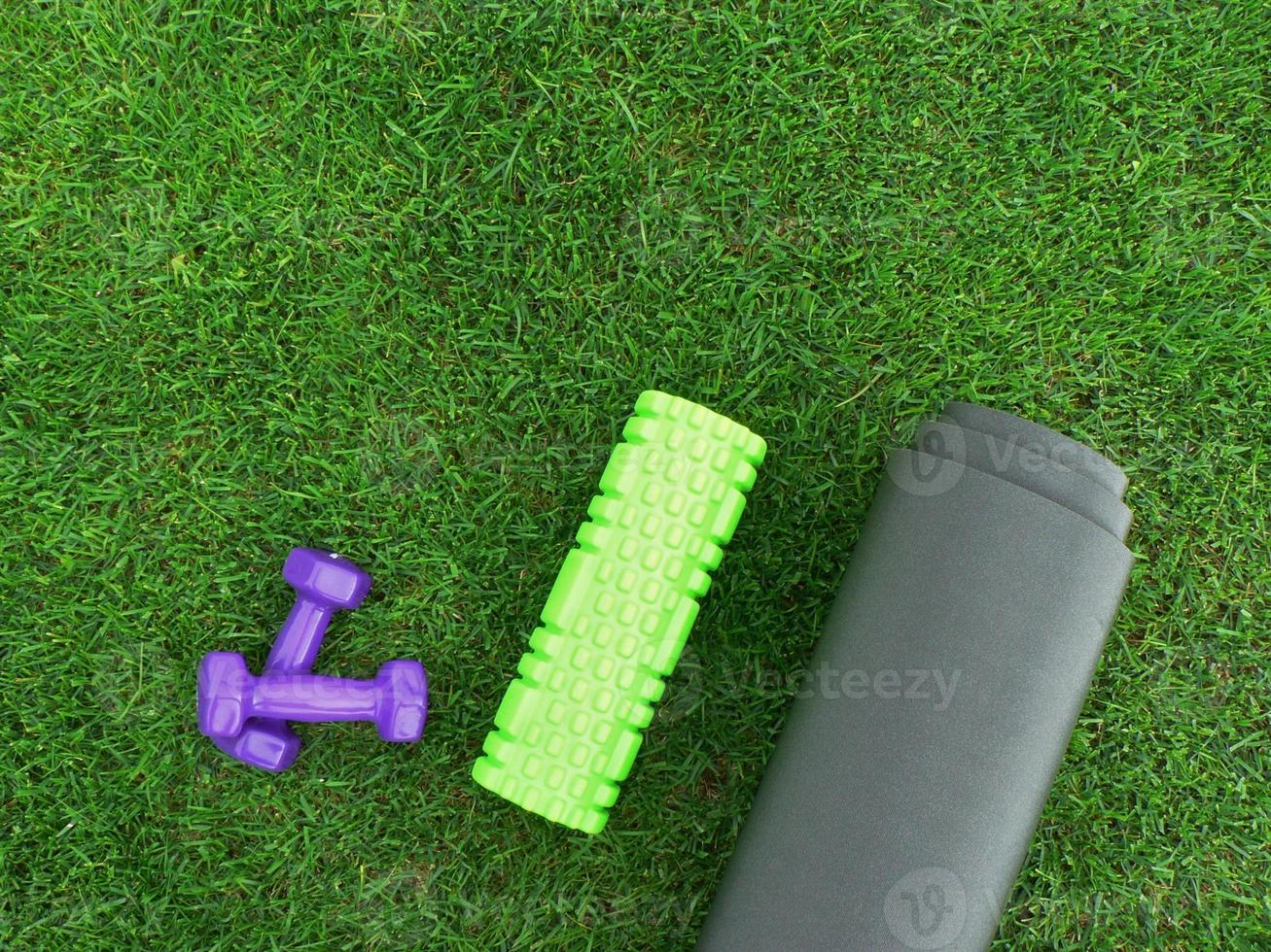 Keeping fit and exercising outdoor or at home. Purple dumbbells, green foam roll on a yoga mat on green grass lawn in a backyard or park. Healthy lifestyle. Copy space. photo