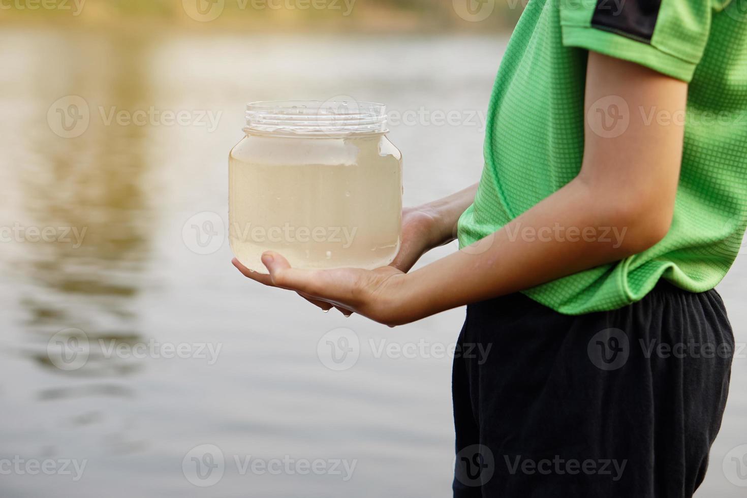 Closeup boy hands holds transparent jar of water at lake. Concept, explore water from nature source.Ecology study. Sample water for examine. Learning outdoor for students about environment. photo