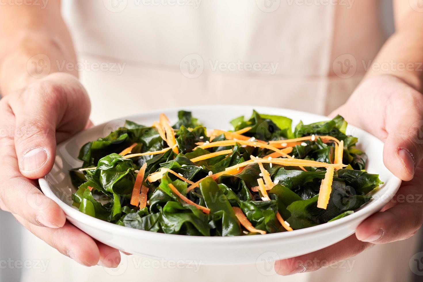 concept of a healthy man wearing an apron with a hand holding a white dish of seaweed salad or kelp on white background photo