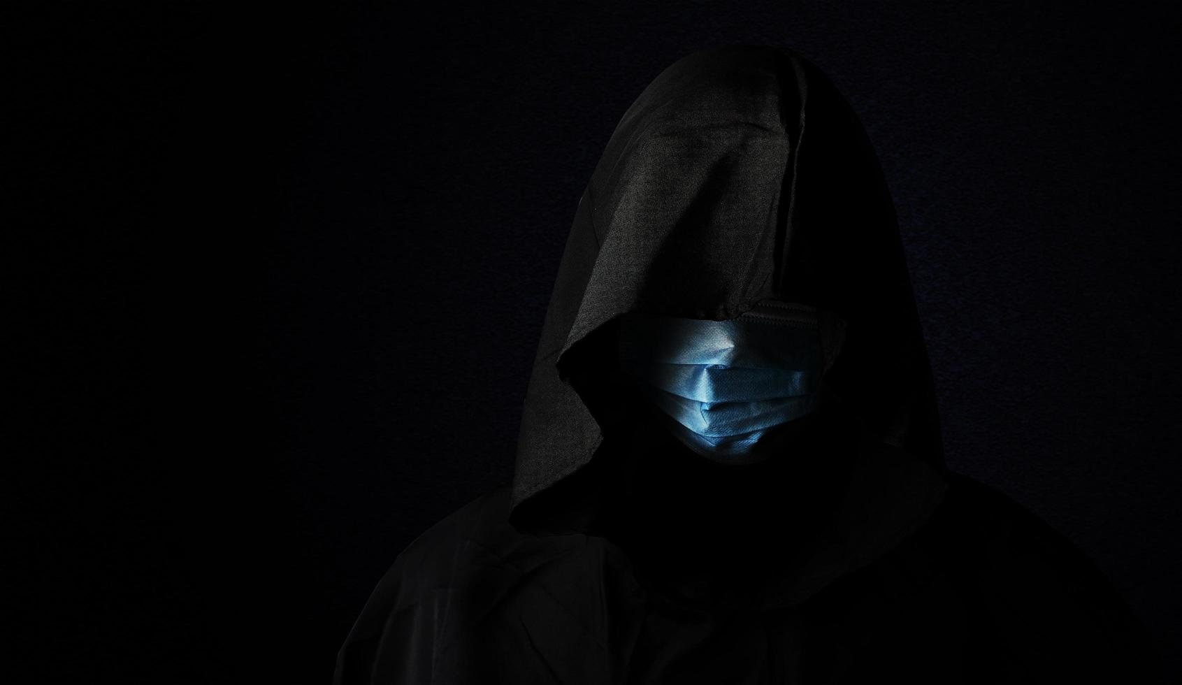 concept of the reaper who wears a medical or surgical mask for protecting corona virus isolated on dark background with clipping path. the man wears a black hood. people photo