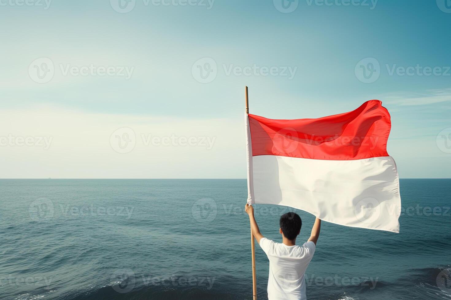 A man holding a red and white Indonesia flag looking at the ocean. photo