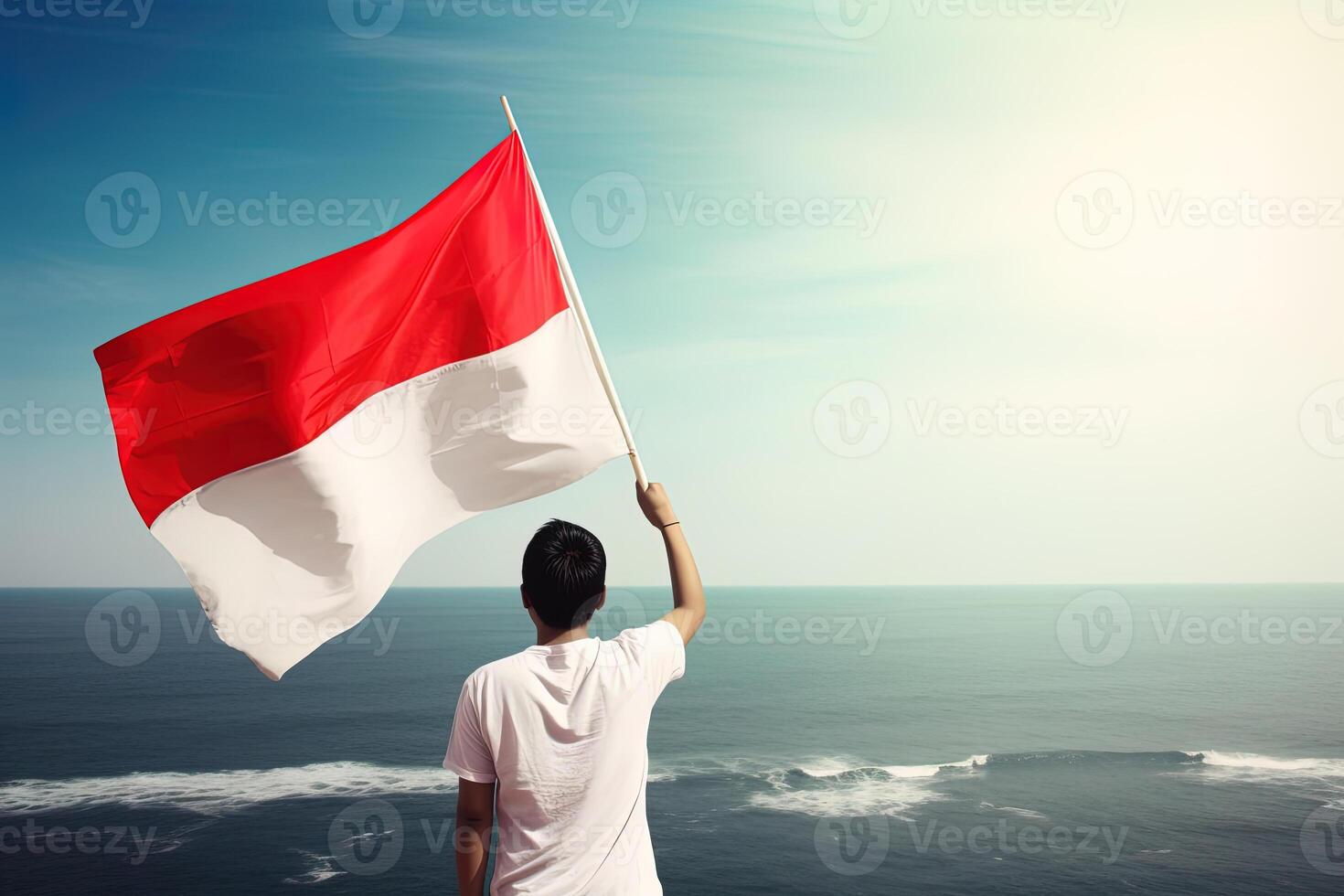 A man holding a red and white Indonesia flag looking at the ocean. photo