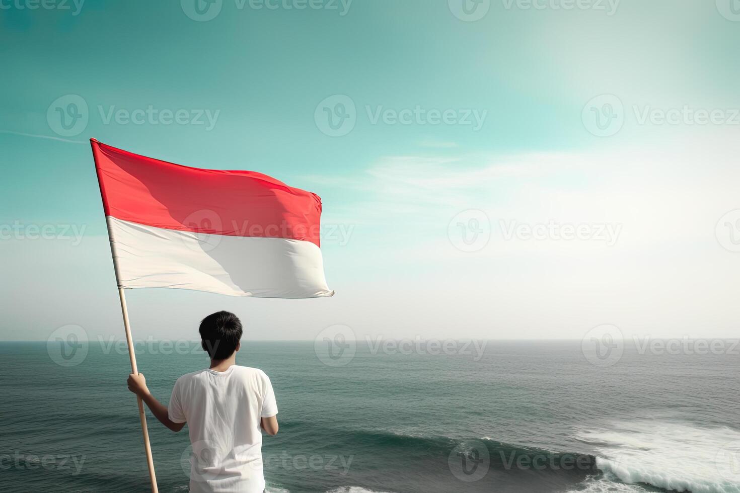 A man holding a red and white Indonesia flag looking at the ocean. photo
