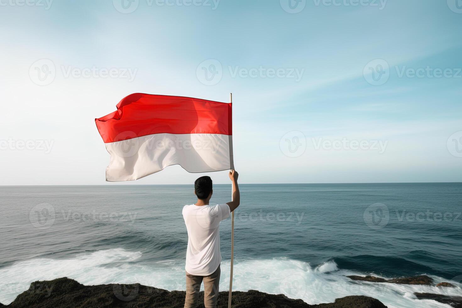 A man holding a red and white Indonesia flag looking at the ocean. photo