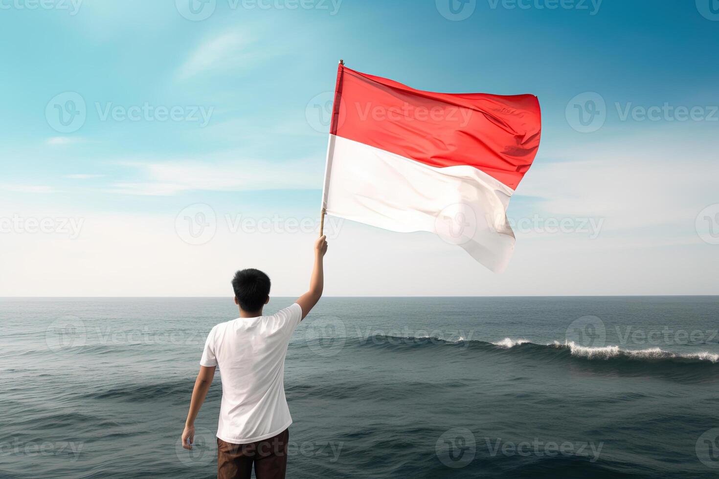 A man holding a red and white Indonesia flag looking at the ocean. photo
