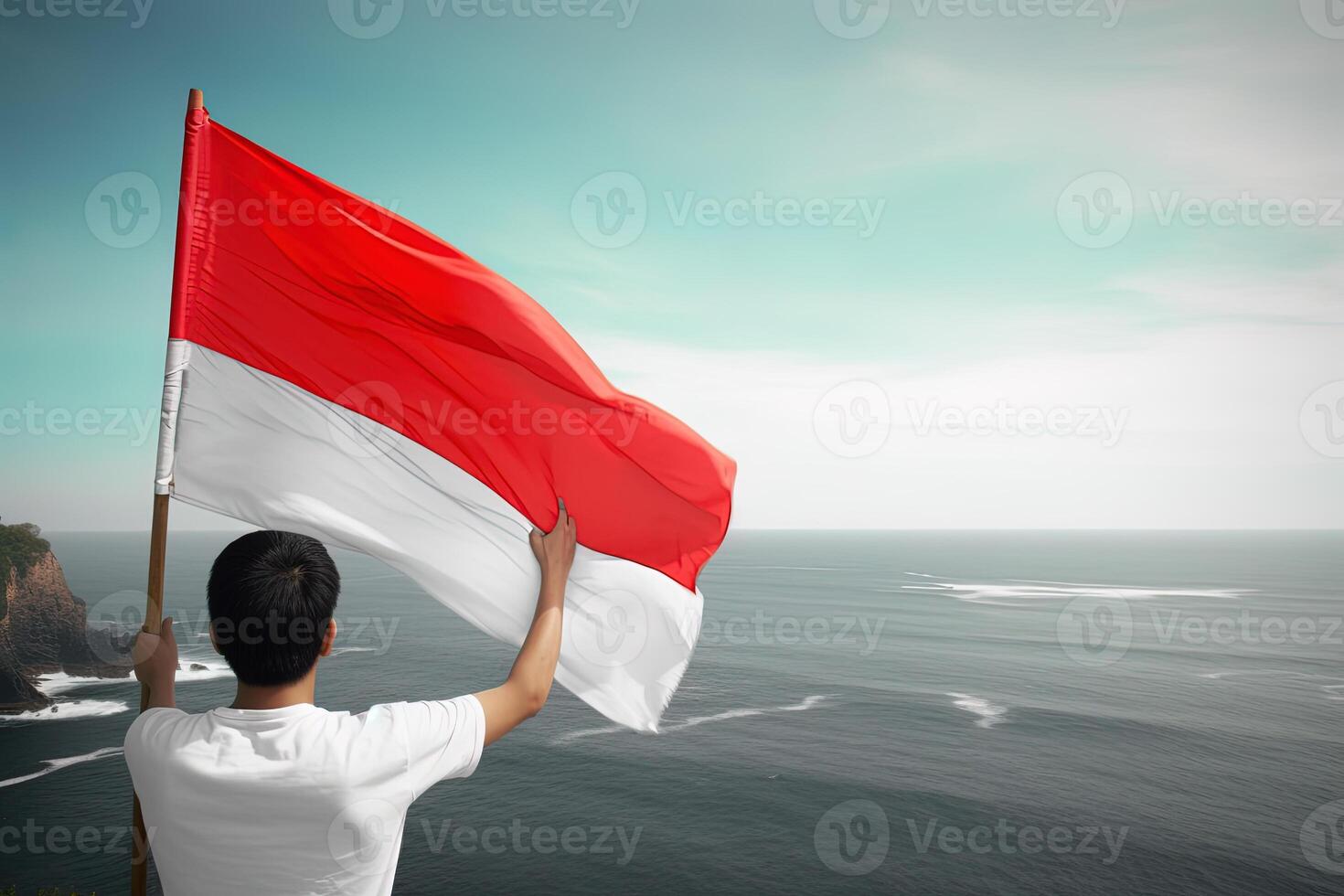 A man holding a red and white Indonesia flag looking at the ocean. photo
