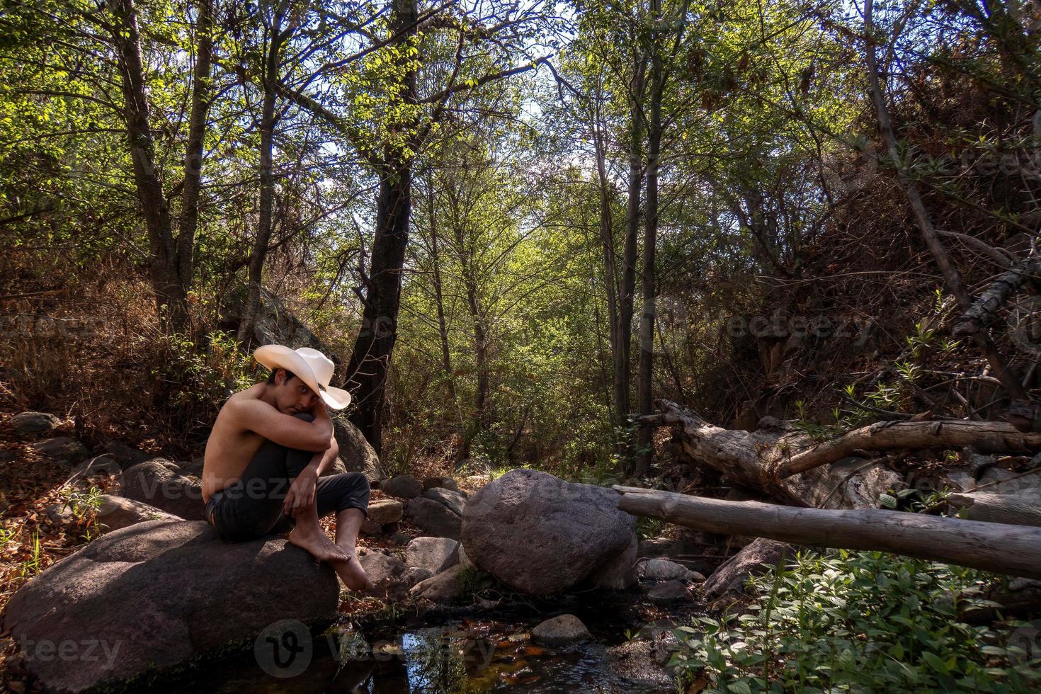 Man sits on a rock in a forest, wearing a cowboy hat. photo
