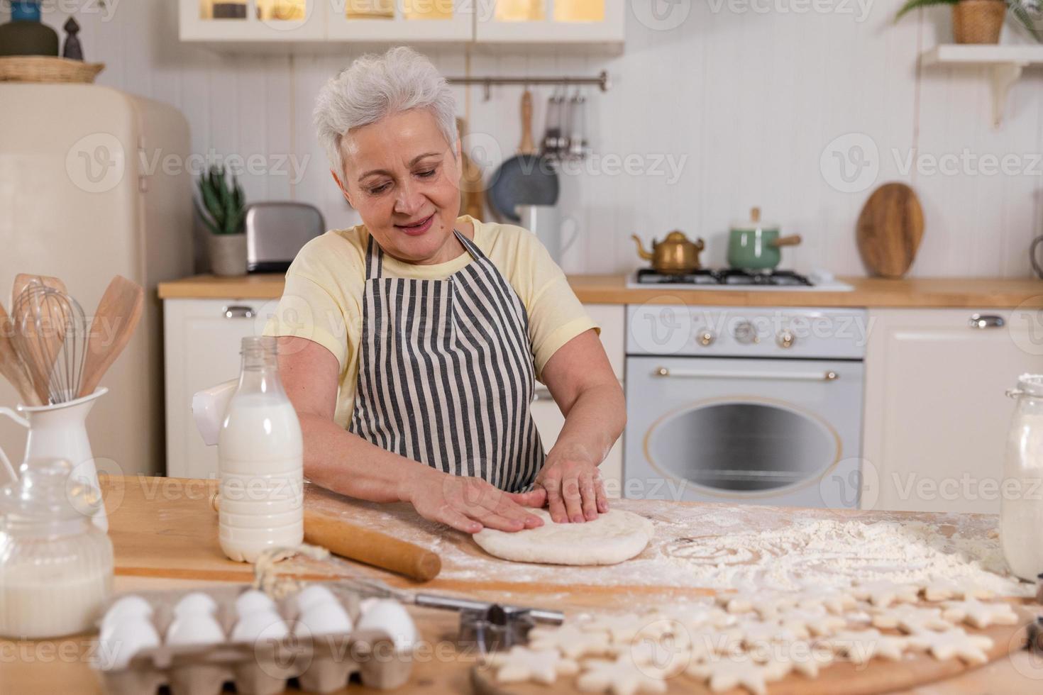 Happy senior woman cooking in kitchen. Stylish older mature gray haired lady grandmother knead dough bake cookies. Old grandma cook homemade food. Household housewife housework concept. photo