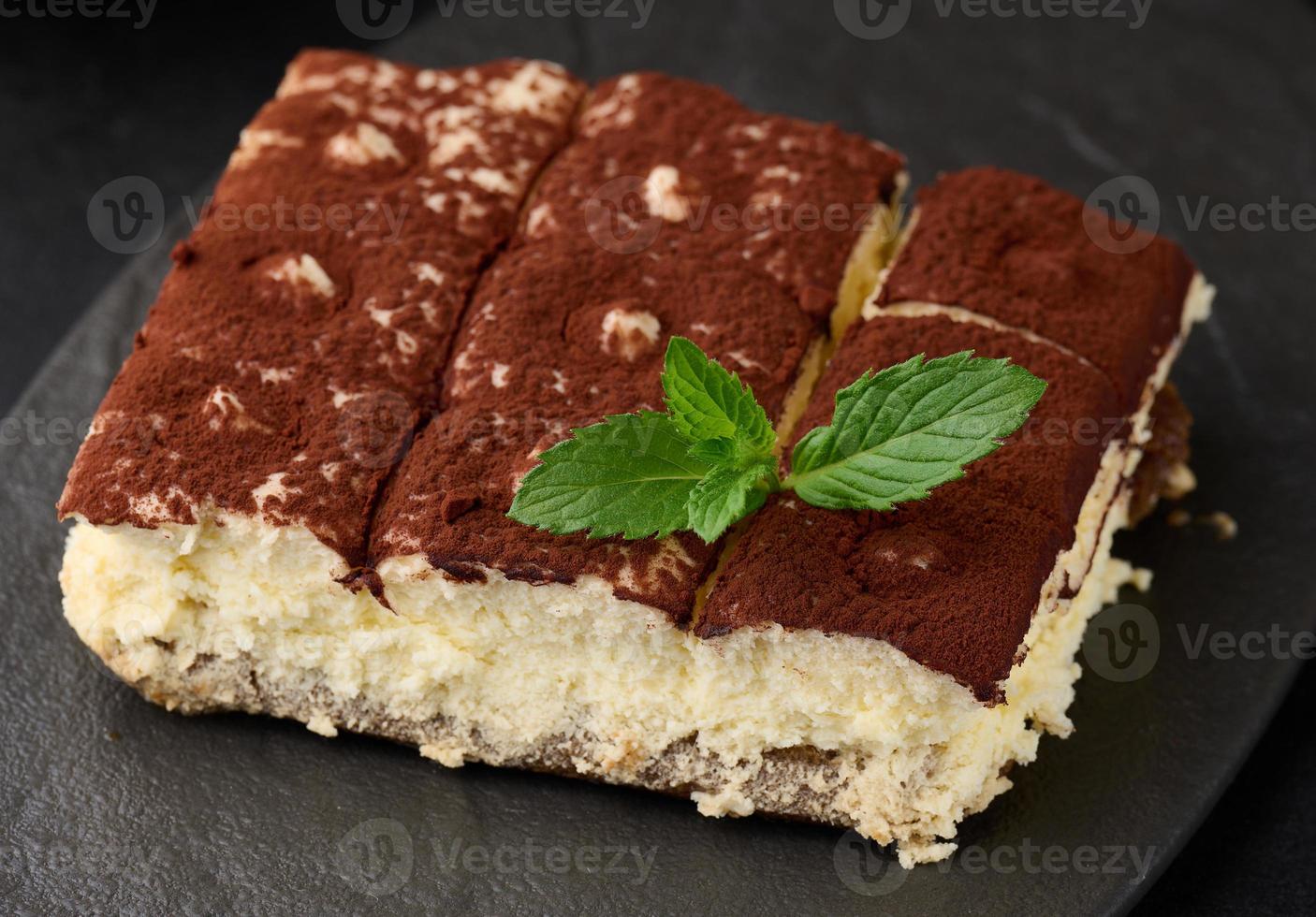 A piece of tiramisu sprinkled with cocoa, on top of a sprig of fresh mint on a black background photo