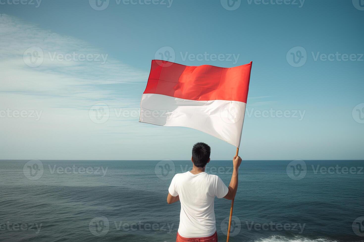 A man holding a red and white Indonesia flag looking at the ocean. photo