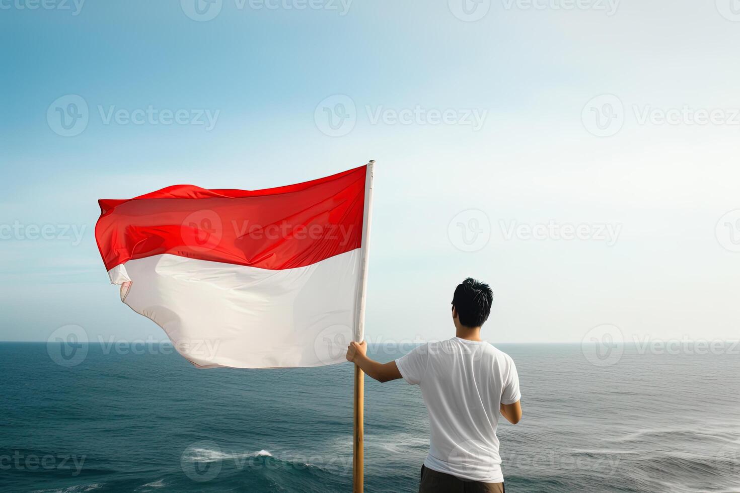 A man holding a red and white Indonesia flag looking at the ocean. photo