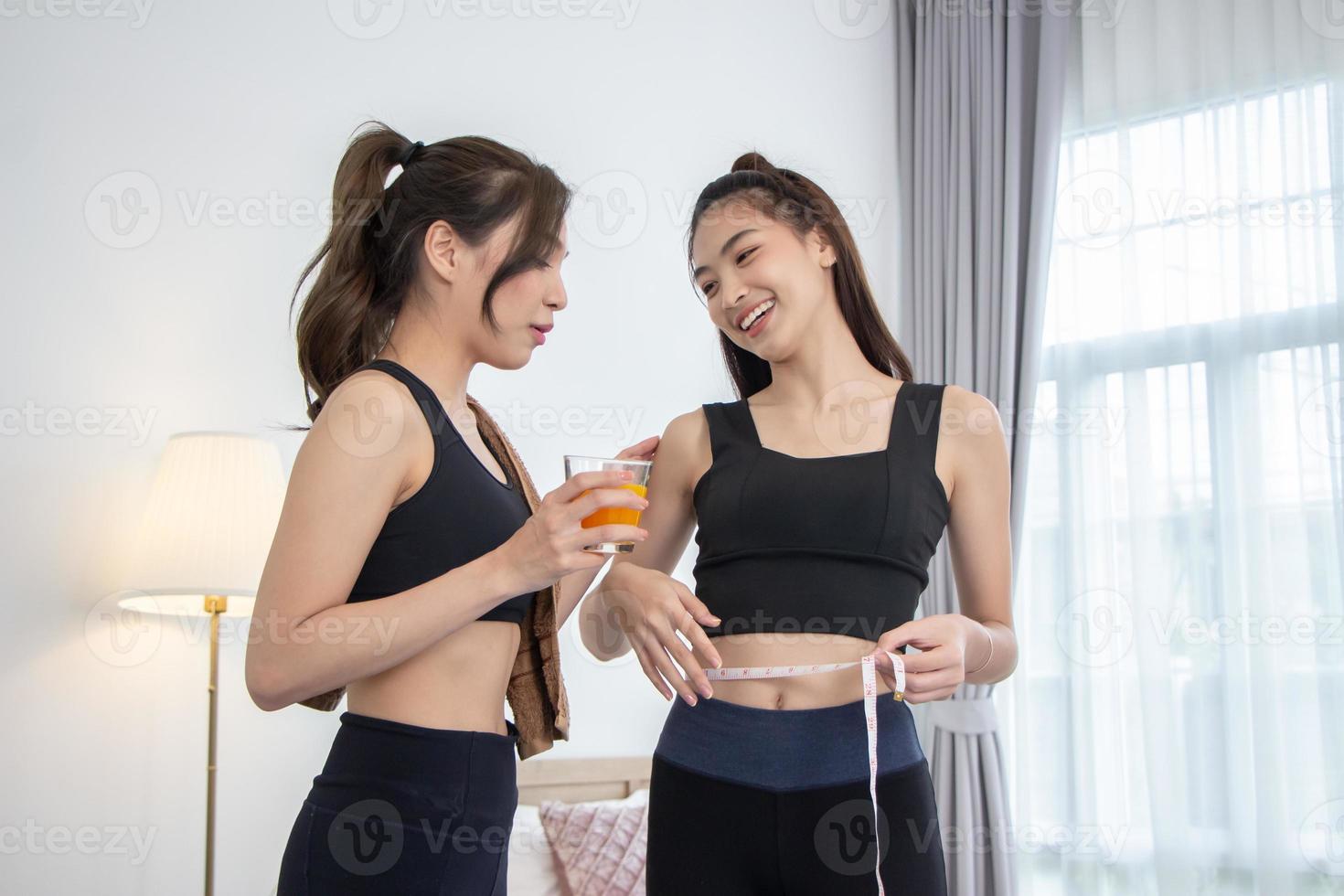 Asian female friends exercising at home. Young woman in workout clothes holding a glass of orange juice and measuring her waist with a tape measure photo