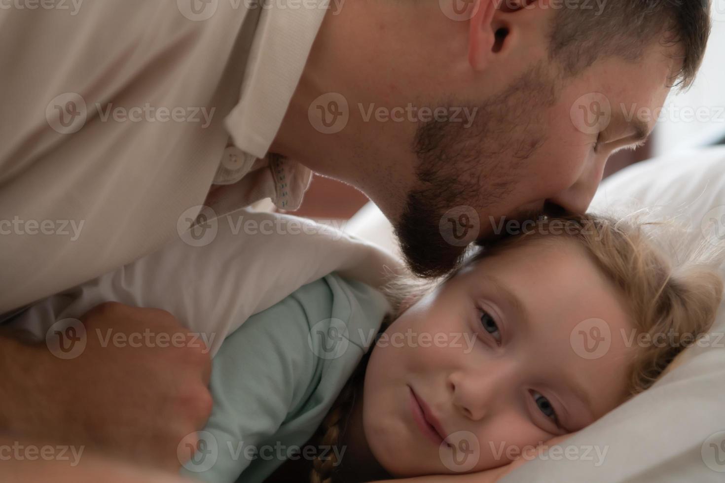 antes de saliente a noche, un padre expresa su afecto por besos su hija en el frente. a obtener un bueno de la noche descanso foto