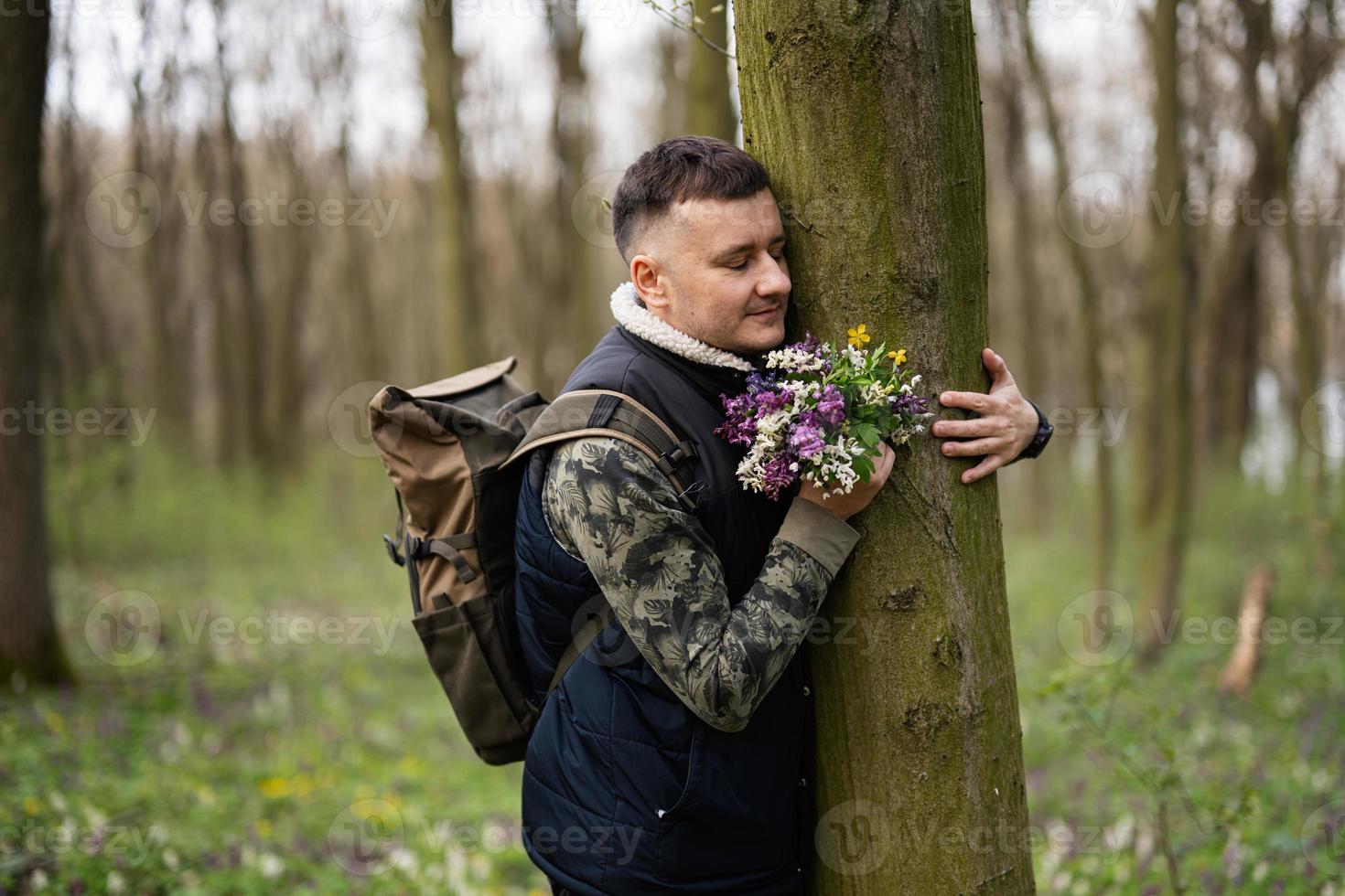 Portrait of man with backpack holding bouquet of spring flowers and hug tree on forest. Love to nature. photo