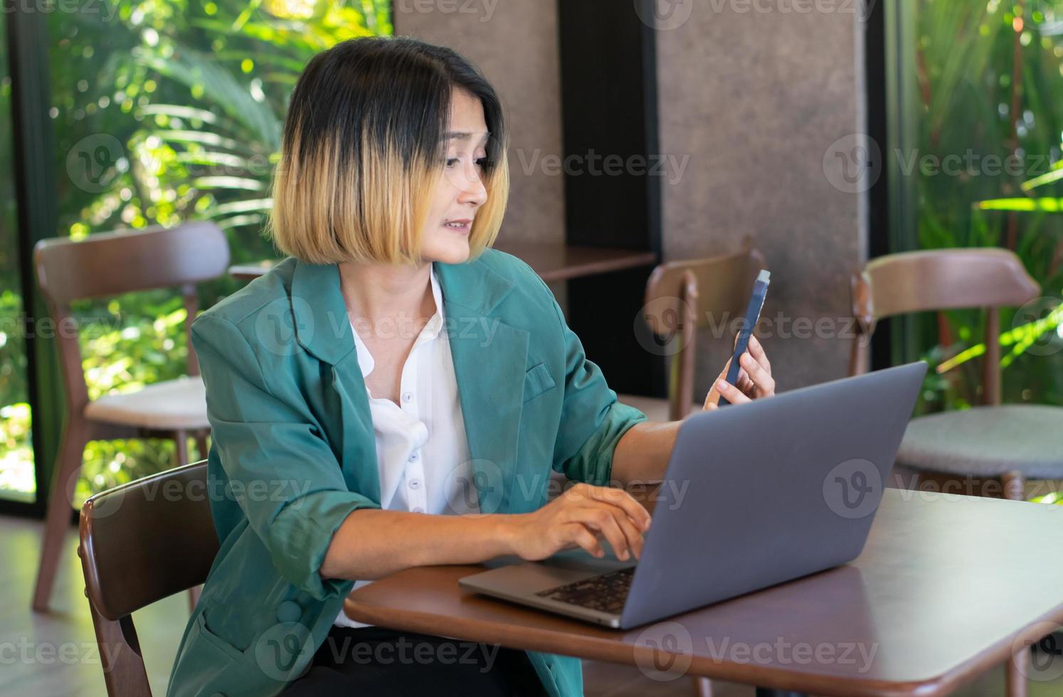 mujer de negocios Lanza libre vistiendo un traje verde sentado en un café y participación móvil teléfono vídeo llamada con cliente y mecanografía en un ordenador portátil. foto