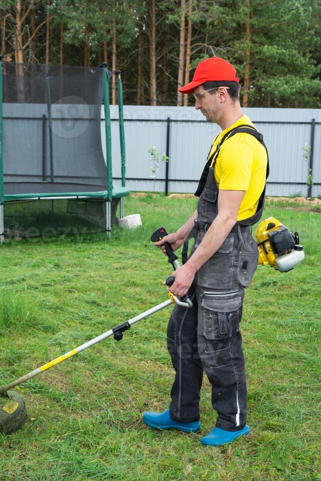 A male gardener mows the green grass of the lawn in the backyard with a gasoline mower. Trimmer for the care of a garden plot photo