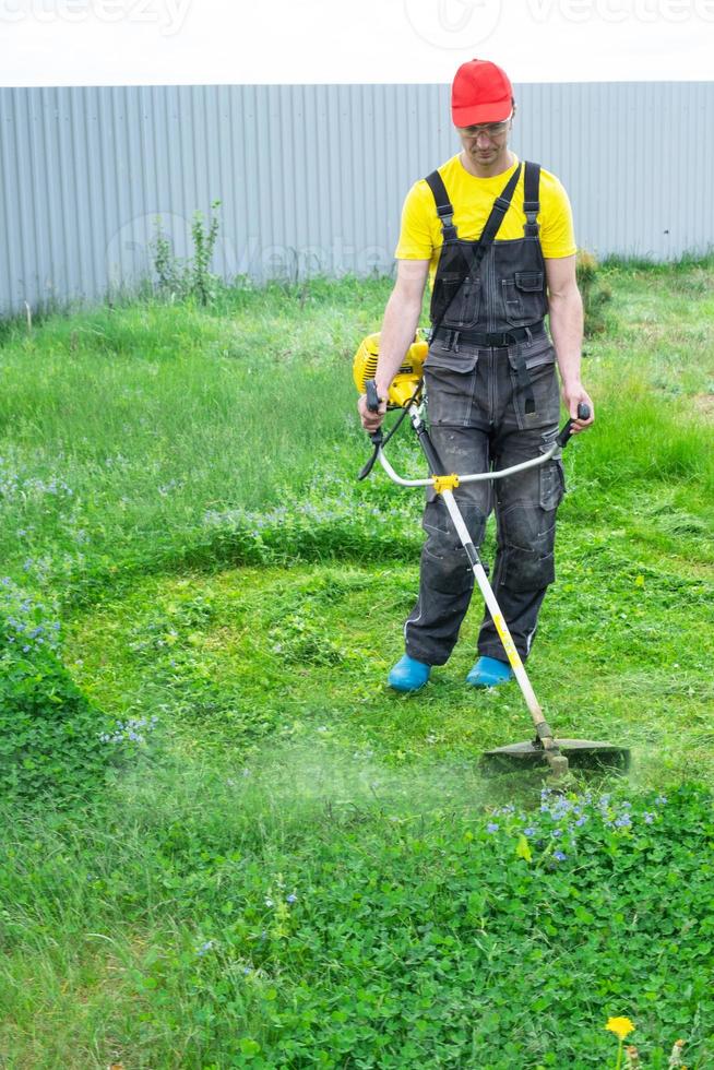 A male gardener mows the green grass of the lawn in the backyard with a gasoline mower. Trimmer for the care of a garden plot photo