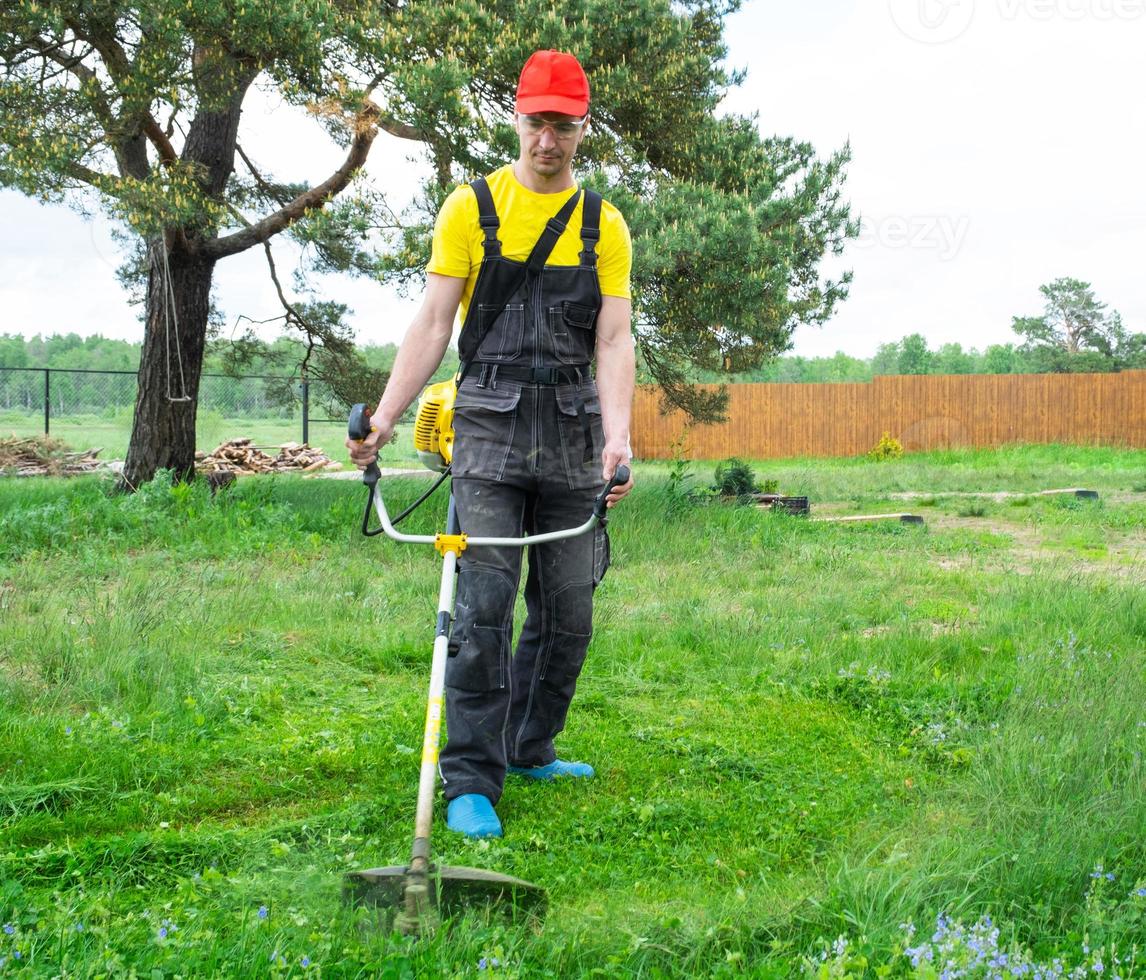 A male gardener mows the green grass of the lawn in the backyard with a gasoline mower. Trimmer for the care of a garden plot photo