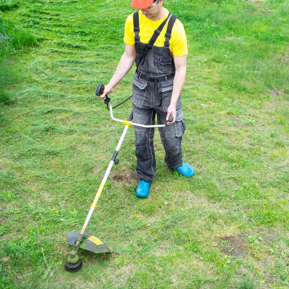 A male gardener mows the green grass of the lawn in the backyard with a gasoline mower. Trimmer for the care of a garden plot photo