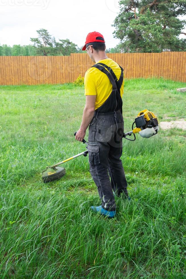 A male gardener mows the green grass of the lawn in the backyard with a gasoline mower. Trimmer for the care of a garden plot photo