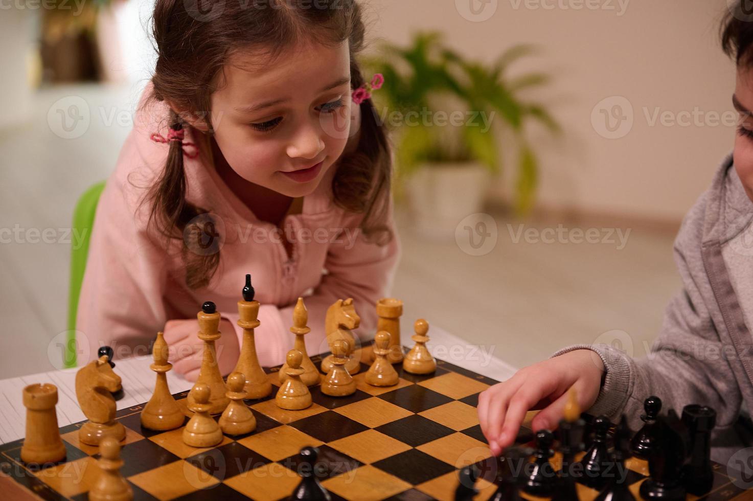 de cerca retrato de un hermosa linda pequeño europeo niña jugando ajedrez juego con su mayor hermano quien escoge arriba un ajedrez pedazo y hacer su mover. intelectual inteligente desarrollo tablero juegos para niños foto