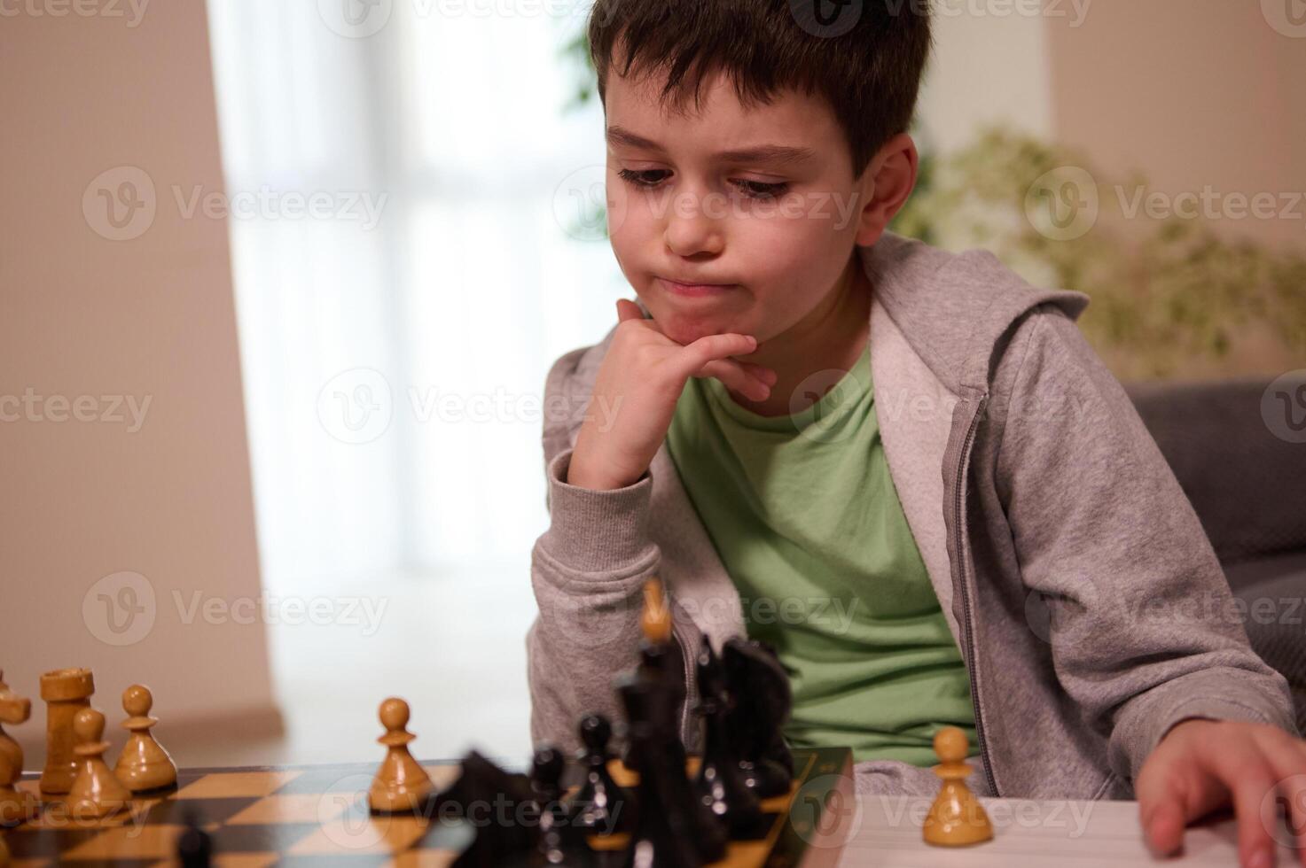 Thoughtful boy of school age keeps his hand on his chin, looks thoughtfully at the chessboard,developing game strategy and moves in a chess game photo