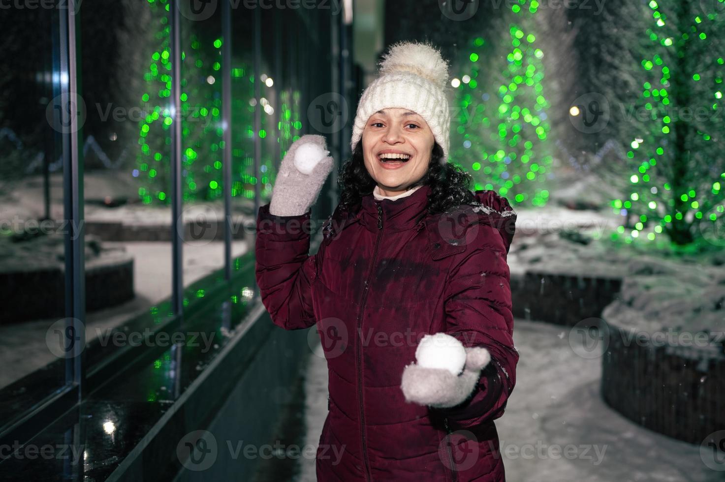Pretty woman plays with snowballs on winter snowy evening, on street lightened by garlands. Winter fun - snowball fight photo