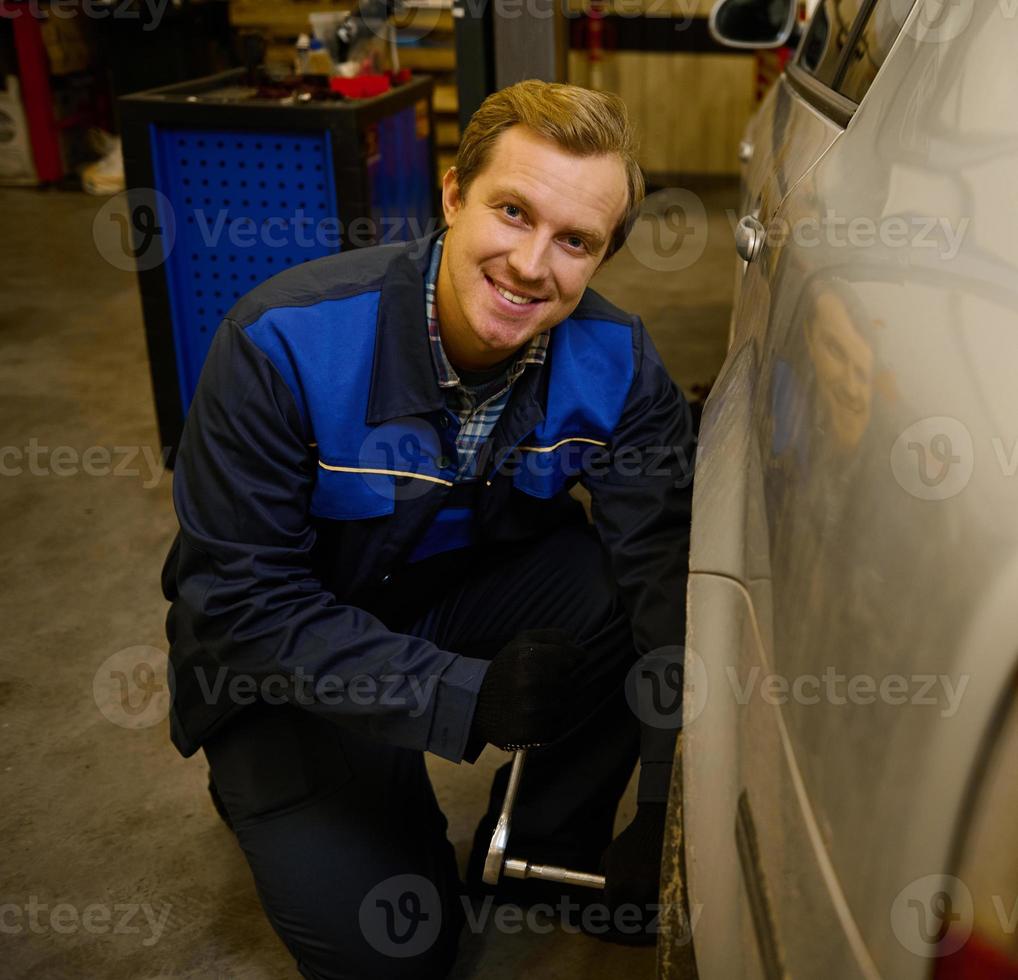 Handsome Caucasian man, professional auto mechanic manually changes the wheel in the car, unscrewing the nuts and bolts with the wrench, smiling at the camera in the repair shop photo