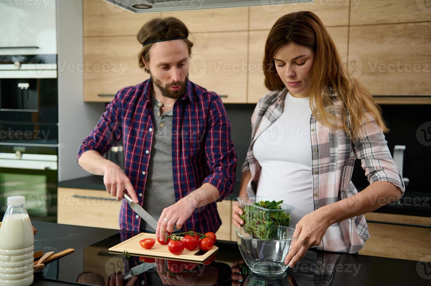 Pregnant woman holds herbs and puts them into a bowl while her husband cuts tomatoes. Beautiful heterosexual couple cooking together in a modern home kitchen photo