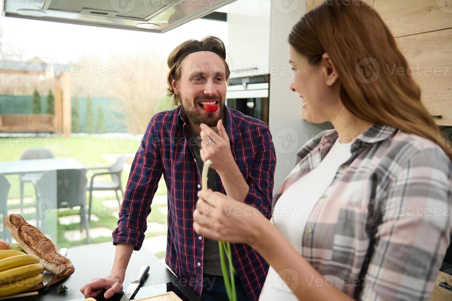 Cooking healthy food. Happy young loving Caucasian couple enjoying time together while cooking in the kitchen while man eating tomato halves and looking at his beloved pregnant woman photo