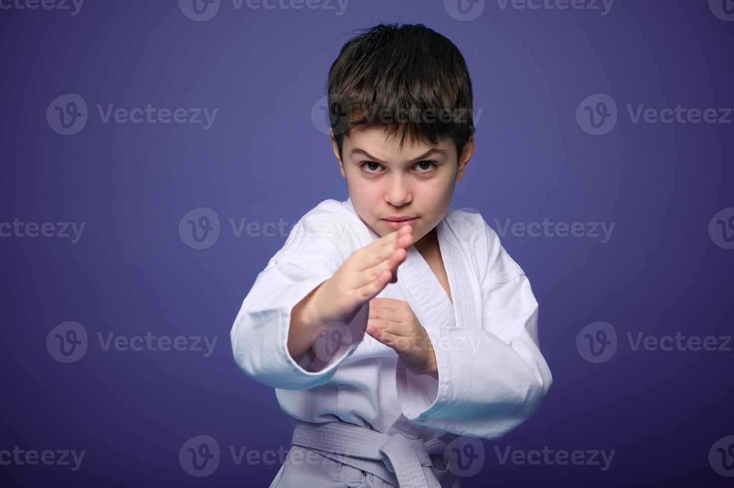 Confident strong kid, aikido fighter practicing martial skills on purple background with advertising copy space. Oriental martial arts practice concept photo