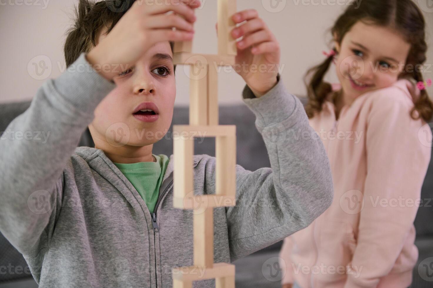 Handsome schoolboy, concentrated on construction of complex tall structure from wooden blocks, against the background of his cheerful little sister, smiling sweetly, looking at his structure. Close-up photo