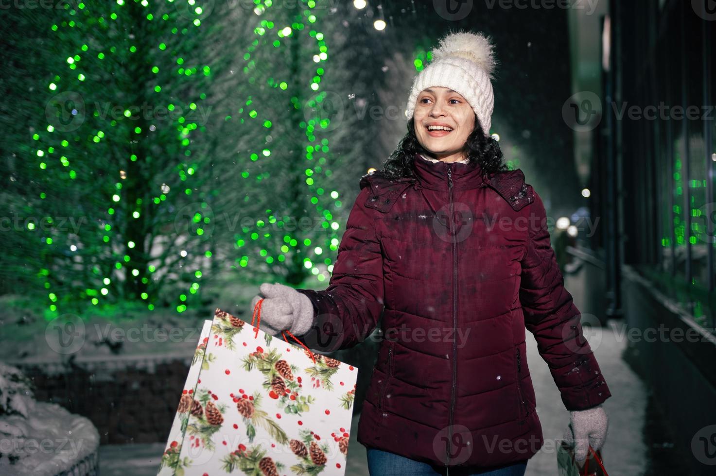 bonito mujer caminando con compras pantalones en el calle iluminado por guirnaldas a Nevado invierno noche foto