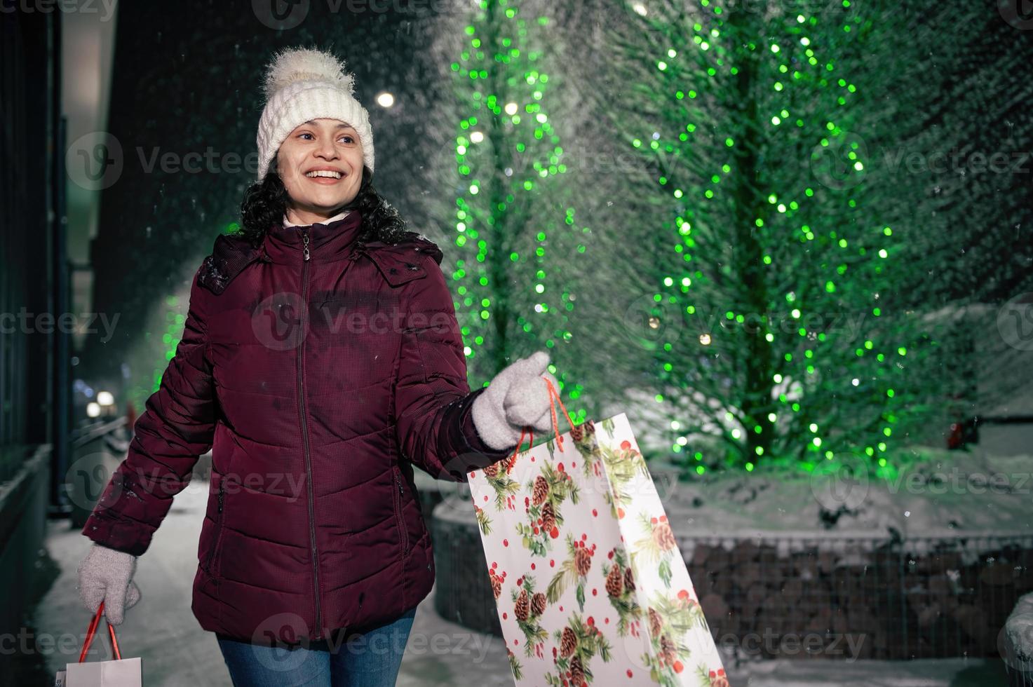 encantador Hispano mujer camina con compras pantalones en un ciudad calle iluminado por guirnaldas a Nevado invierno noche foto