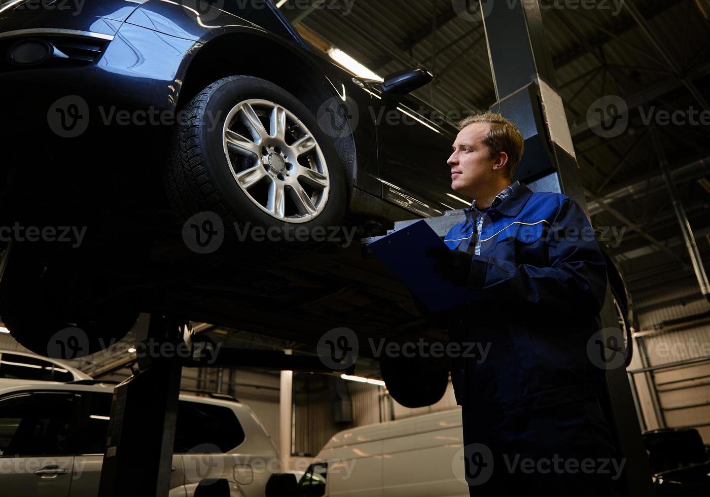 Portrait of a mechanic, technician, car engineer in uniform making the checklist on clipboard for repairing the car lifted on a hoist in the repair shop during warranty car maintenance. Auto service photo