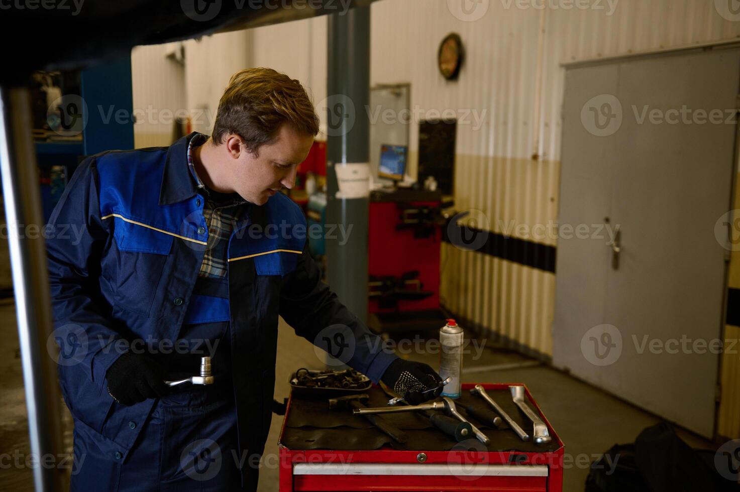 Auto mechanic in uniform with work tools, inspecting a lifted car on a hoist, while doing checklist for warranty repairing automobile in the workshop garage photo