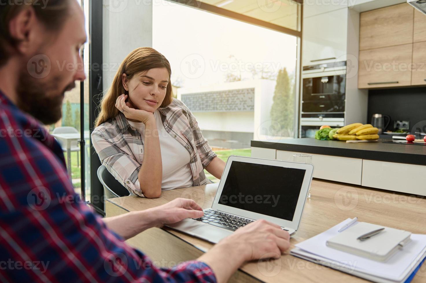 Rear view of a woman looking at the laptop screen monitor of her husband working remotely from home. Stack of documents and notepad lying down on the desk. Copy space for text photo