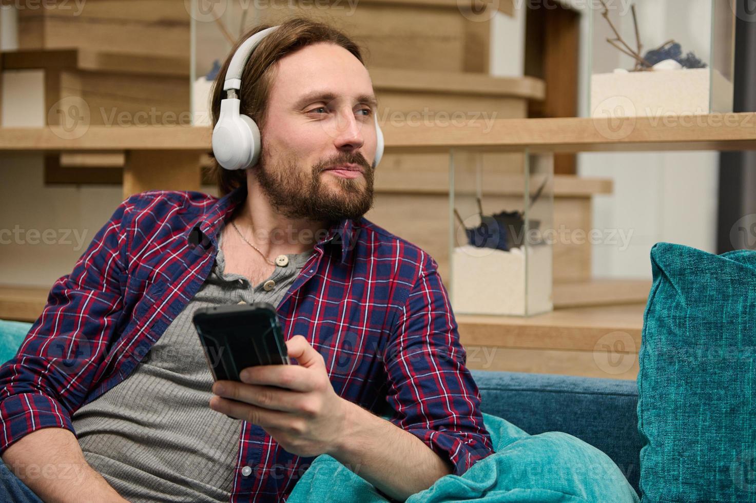 medio Envejecido caucásico barbado hermoso hombre con inalámbrico auriculares, sostiene un teléfono inteligente en su mano y soñando mirando aparte foto