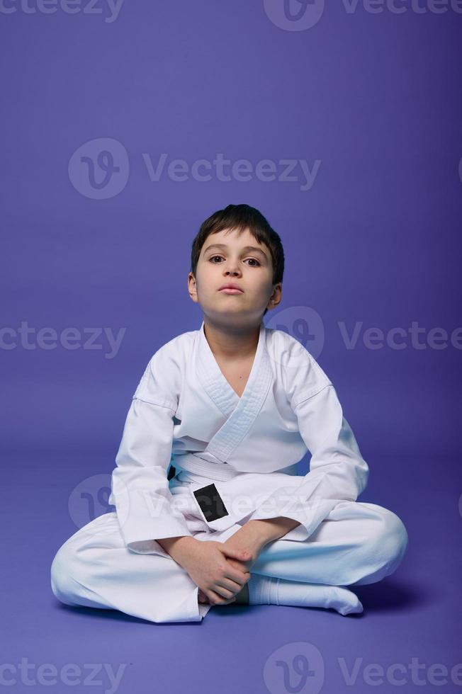 Confident European teenager - aikido fighter - in white kimono sitting in lotus pose while practicing oriental martial arts on purple background with copy space photo