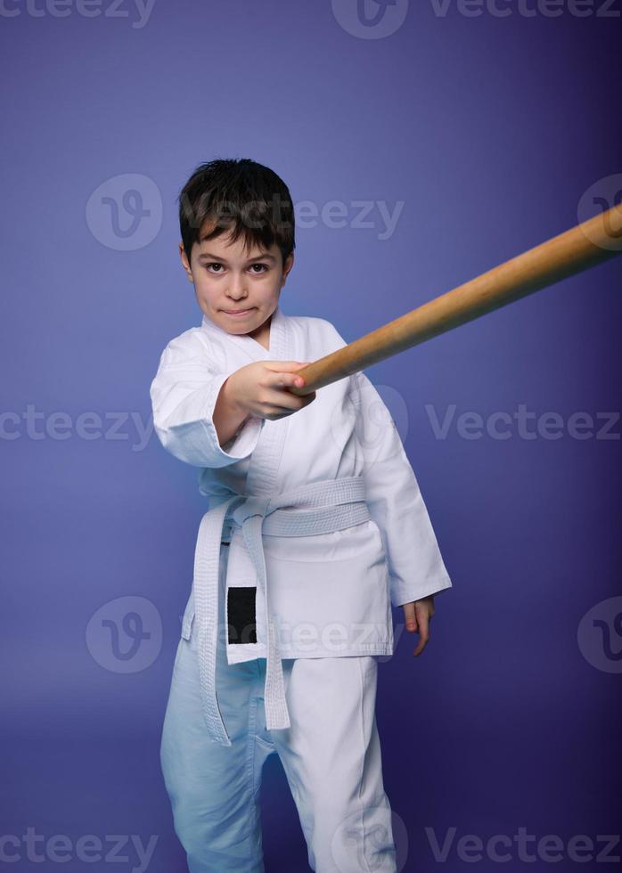 Oriental martial arts practice, combat, aikido, japanese sports and philosophy. Child boy fighter with wooden weapon bokken sword on violet background photo