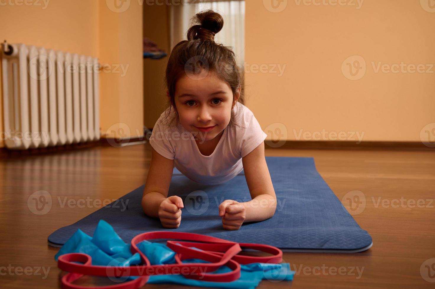 Determined sporty little girl doing plank exercise on a blue fitness mat while working out and stretching body indoors photo