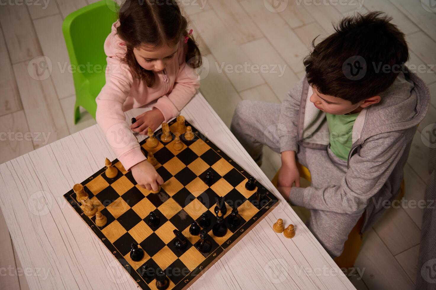 Overhead view of a cute little girl sitting at the table and plays chess with her brother, picking up a chess piece and making his move. Early development, home educational games for children photo